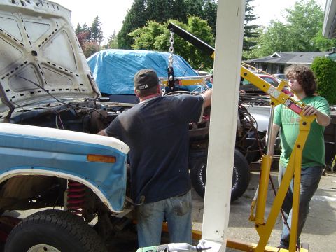 1979 Ford Bronco - Two men work on a 1979 Ford Bronco in a driveway, with another vehicle covered in a blue tarp behind it.