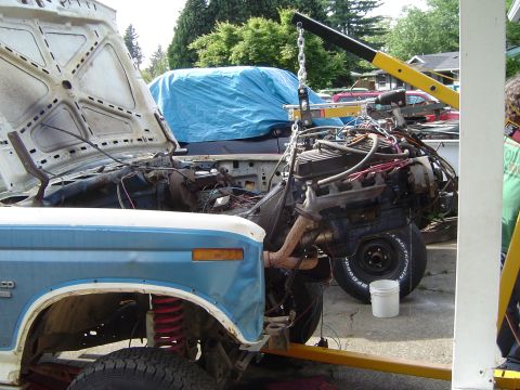 1979 Ford Bronco - An engine is being hoisted out of a blue and white SUV, with another vehicle covered in a blue tarp in the background.