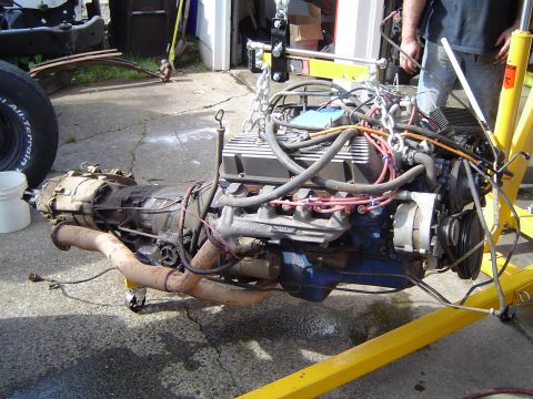 1979 Ford Bronco - Engine and transmission assembly suspended by a yellow engine hoist, with a partial vehicle frame in the background.
