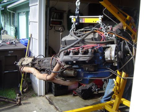 1979 Ford Bronco - Engine and transmission assembly removed from a vehicle and suspended by a yellow engine hoist in a garage setting.