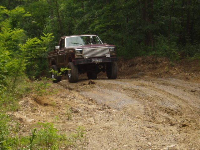 1980 Ford Bronco driving on a muddy dirt road in a wooded area
