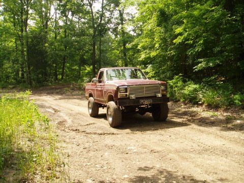 1980 Ford Bronco - A maroon pickup truck drives on a dirt road through a wooded area.