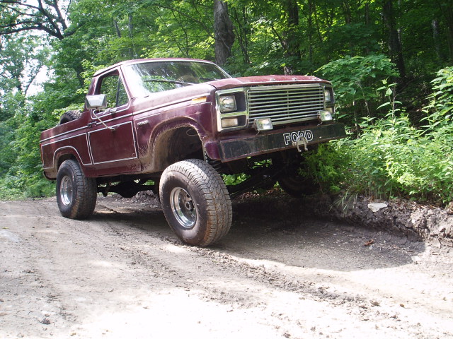 Maroon Ford Bronco with lifted suspension driving on a dirt trail, front grille visible