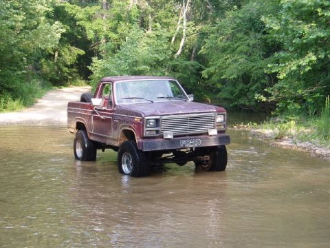 1980 Ford Bronco - This maroon Ford F-Series pickup truck is navigating a shallow creek in a wooded area.