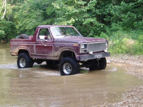 1980 Ford Bronco - This lifted Ford Bronco is navigating a water crossing surrounded by trees.