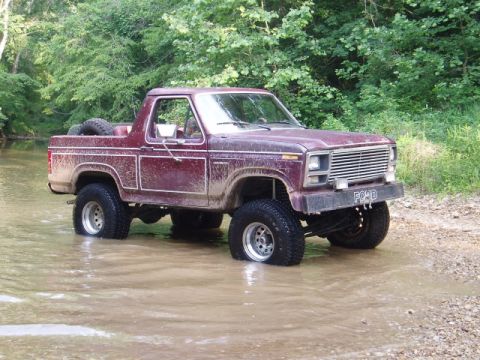 1980 Ford Bronco - A maroon Ford Bronco with white trim is driven through a shallow stream, its tires partially submerged.