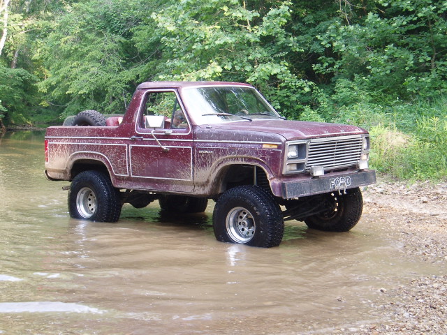 Maroon Ford Bronco with white trim driving through shallow water, spare tire mounted in the bed
