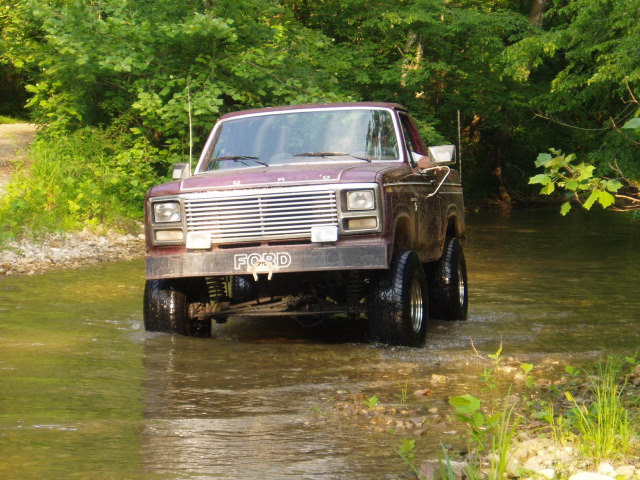 1980 Ford Bronco driving through shallow water with trees in background