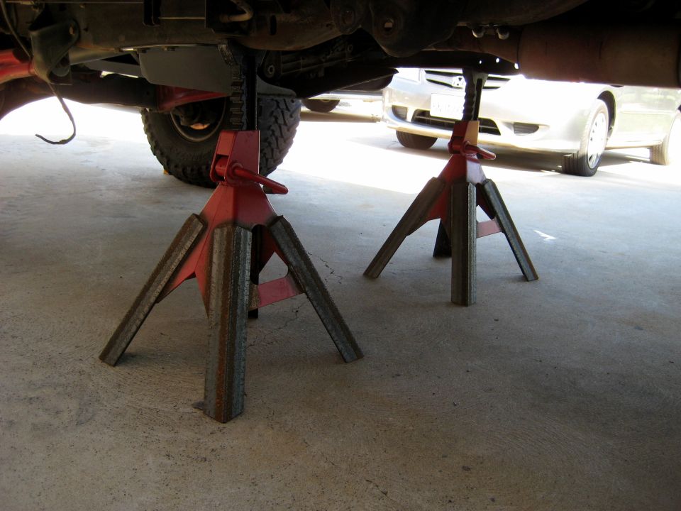 Undercarriage of a vehicle supported by red jack stands, with another vehicle visible in the background