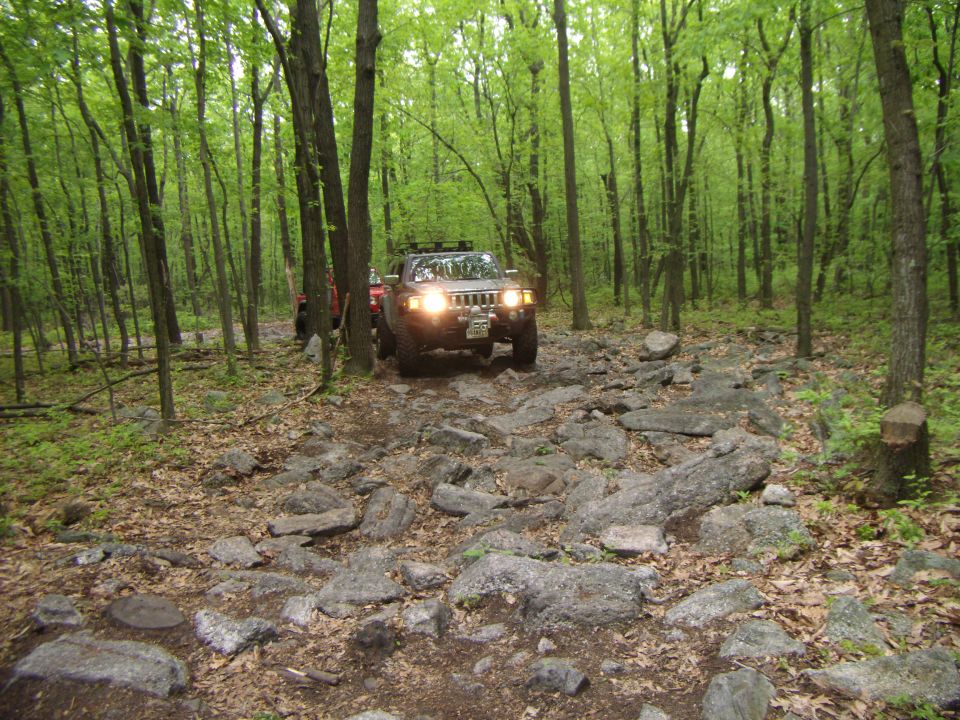 Brown Hummer H3 on rocky forest trail, front grille visible, headlights on