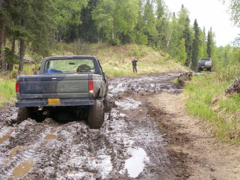 1981 Jeep CJ7 - A camouflaged pickup truck struggles through deep mud on a wooded trail, with another vehicle visible further down the path.