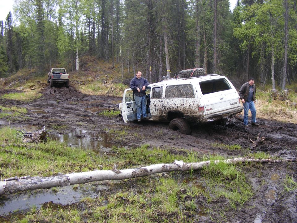 White GMC SUV stuck in mud, another truck in background. Two men standing near SUV
