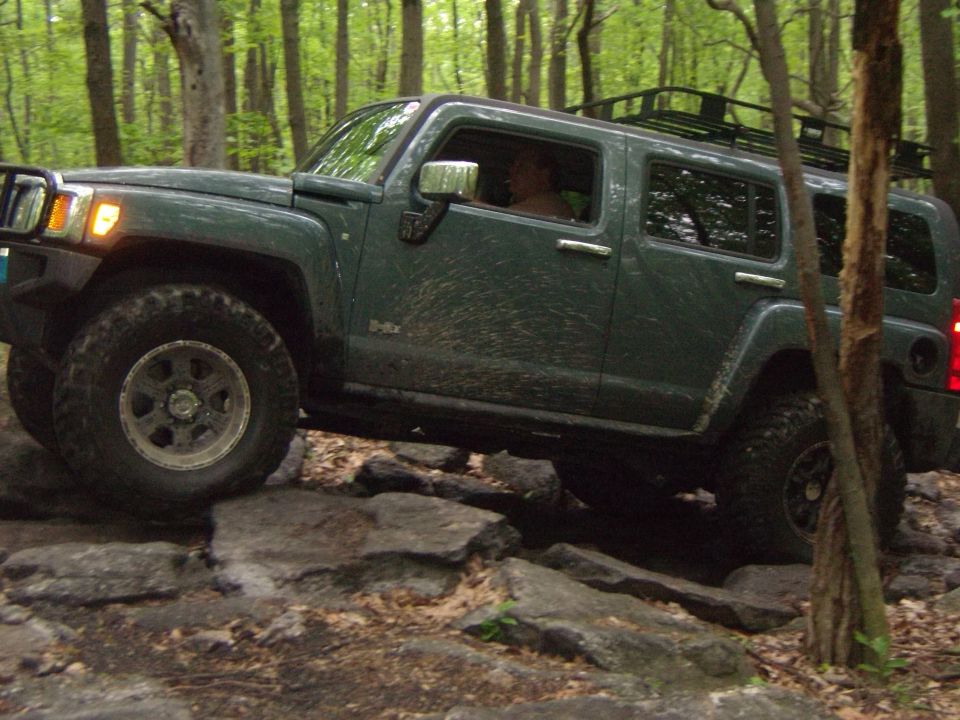 Green Hummer H3 driving over rocks in a wooded area. Has a roof rack