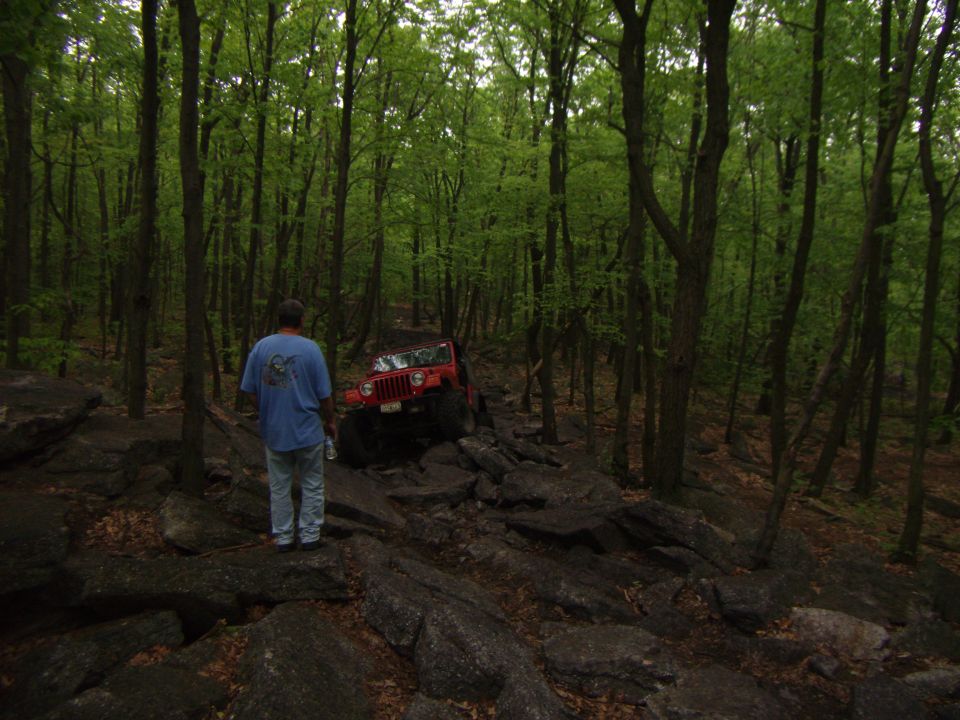 Red Jeep Wrangler on a rocky forest trail with a person standing nearby.