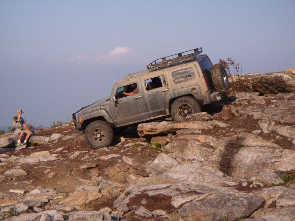 Hummer H3 driving on a rocky hill with a person sitting nearby.