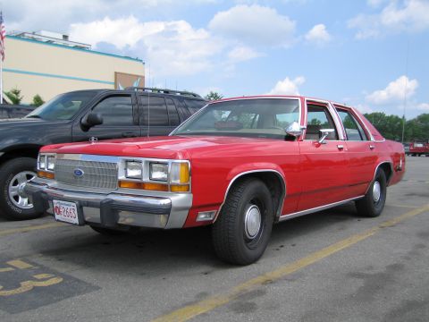 1991 Ford LTD Crown Victoria - red Ford four-door sedan with a Massachusetts plate parked in a lot next to a black SUV under blue sky.