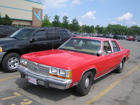 1991 Ford LTD Crown Victoria - Front three-quarter view of a red Ford four-door sedan parked beside a black Ford SUV in a lot.