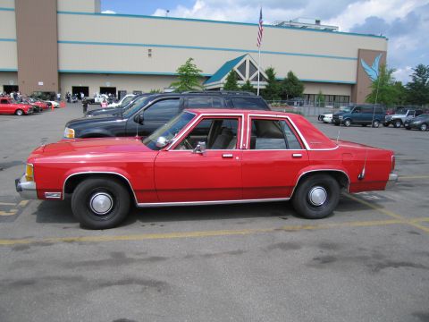 1991 Ford LTD Crown Victoria - Side profile of a red Ford full-size four-door sedan parked in a lot, with an American flag and building behind.