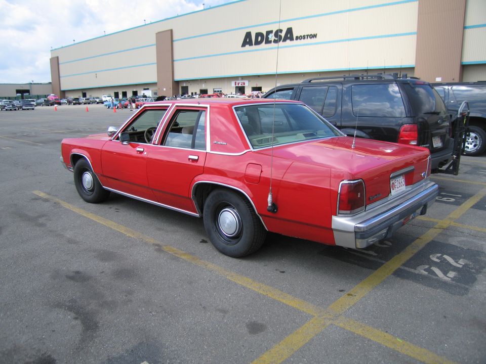 Rear three-quarter view of red Ford sedan in lot, ADESA BOSTON building behind