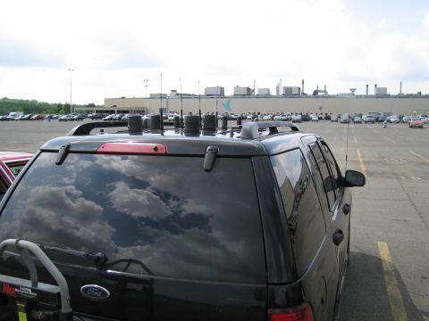 1991 Ford LTD Crown Victoria - Rear of a black Ford SUV fitted with several roof-mounted antennas, parked in a large lot under cloudy sky.