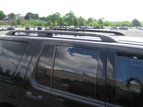 1991 Ford LTD Crown Victoria - Close-up of a black SUV's roof rails and tinted rear side windows reflecting clouds in a parking lot.