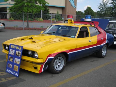 1991 Ford LTD Crown Victoria - yellow, red and blue Mad Max Interceptor replica car with roof lights displayed beside a Mad Max sign.