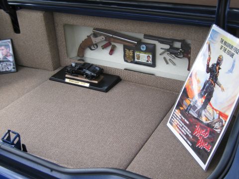 1991 Ford LTD Crown Victoria - carpeted trunk display featuring a model car, a replica sawed-off shotgun and revolver, and a Mad Max movie poster.