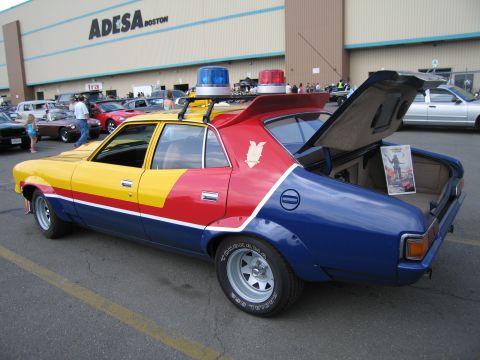 1991 Ford LTD Crown Victoria - Rear three-quarter view of the yellow, red and blue Mad Max Interceptor replica with roof lights and trunk open at a show.