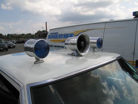 1991 Ford LTD Crown Victoria - Two blue dome beacons and a chrome siren speaker mounted on a white car roof, with a Sheriff van behind.