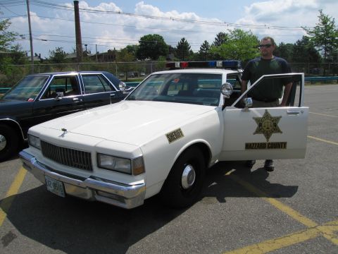 1991 Ford LTD Crown Victoria - man stands at the open door of a white Chevrolet Caprice marked as a Hazzard County Sheriff car in a parking lot.