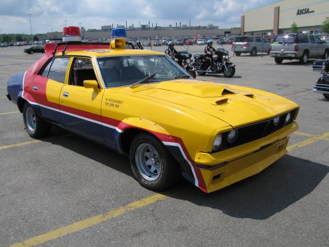 1991 Ford LTD Crown Victoria - yellow and red Ford Falcon pursuit-special replica with roof lights sits in a parking lot near motorcycles.