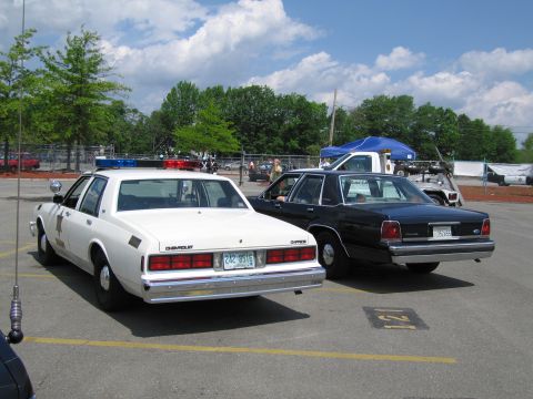 1991 Ford LTD Crown Victoria - white Chevrolet Caprice police car is parked rear-to-rear with a black Ford LTD sedan at an outdoor event.