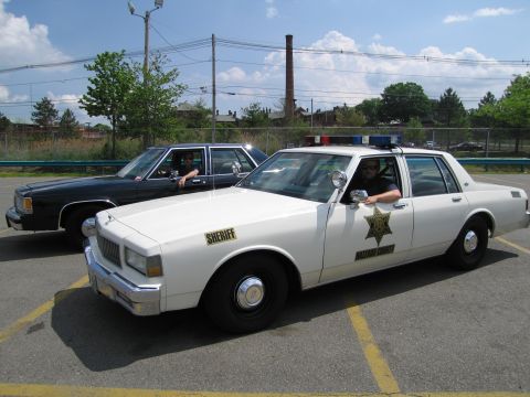 1991 Ford LTD Crown Victoria - white Chevrolet Caprice marked Hazzard County Sheriff with a driver inside is parked ahead of a black sedan.