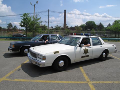 1991 Ford LTD Crown Victoria - white Hazzard County Sheriff Caprice and a black sedan sit in a parking lot, each with a person in the driver seat.