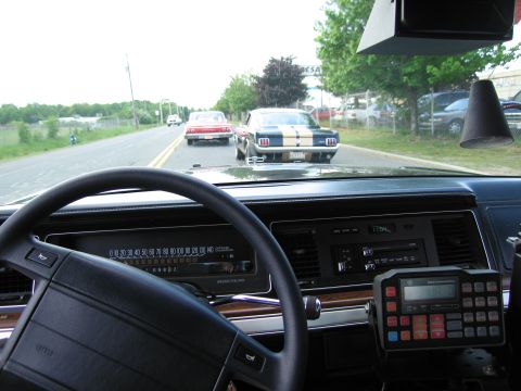 1991 Ford LTD Crown Victoria - Through a windshield, a red and a striped Mustang drive on a road; the dash holds a steering wheel and police radio.