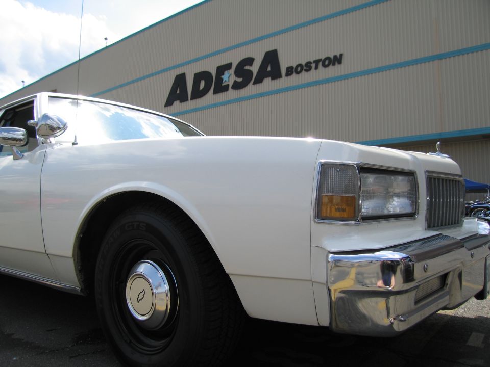 Front corner of a white Chevrolet sedan in front of an ADESA Boston sign