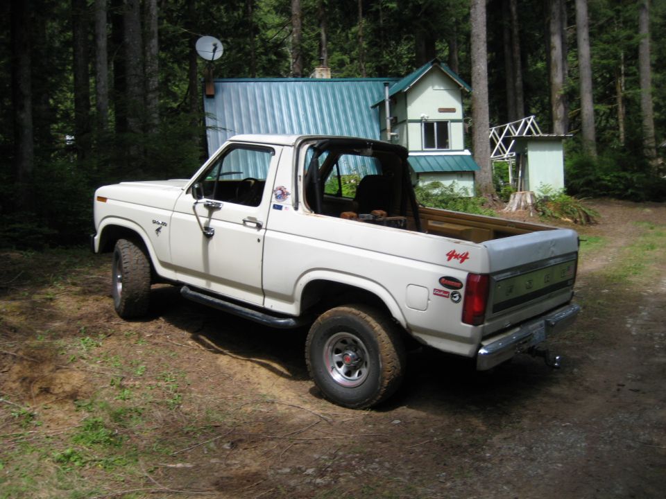 White 1980 Ford Bronco parked in a forest next to a cabin.