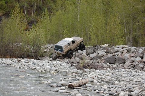 1978 Ford Bronco - A white SUV sits on a rocky shoreline with green foliage in the background.