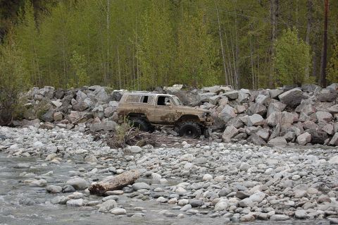 1978 Ford Bronco - A heavily soiled SUV navigates a rocky riverbed, with trees and a rock embankment in the background.