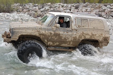1978 Ford Bronco - A driver navigates a heavily muddied SUV through a rocky riverbed, kicking up water.