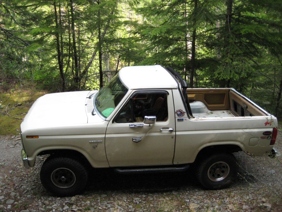 Side view of a cream-colored 1980 Ford Bronco parked in a forest.