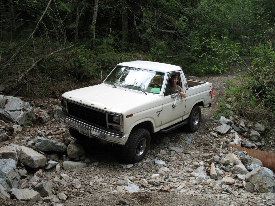 White 1980s Ford Bronco driving on a rocky forest trail.