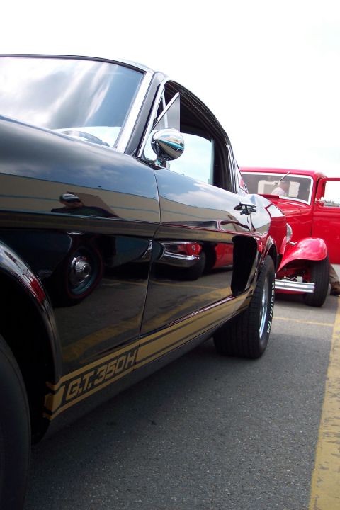 2001 Ford Crown Victoria - A black 1966 Ford Mustang GT350H is parked next to a red hot rod.