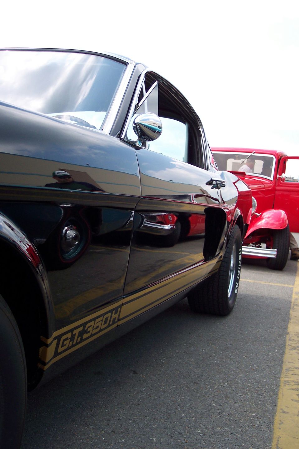 Black 1966 Ford Mustang GT350H side view with gold stripe and a red hot rod behind it