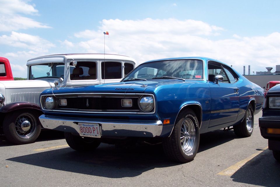 Blue Plymouth Duster parked next to a white sedan and a red truck