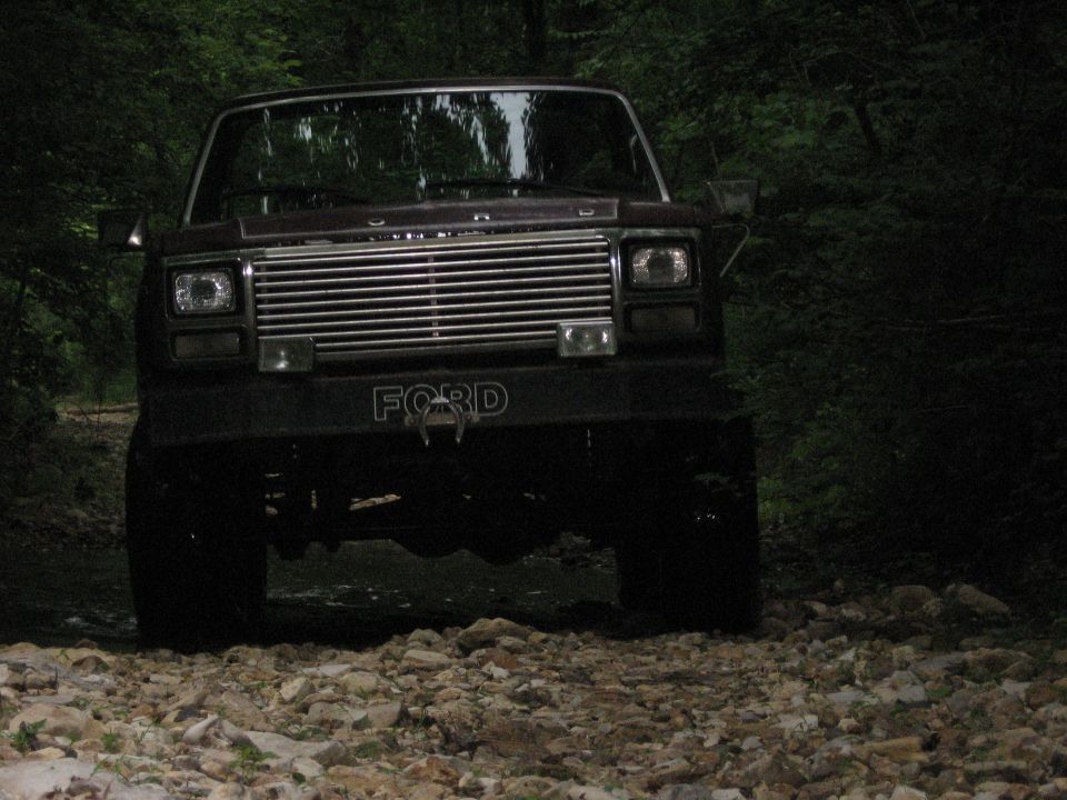 Dark brown 1980 Ford Bronco front view on a rocky path with trees
