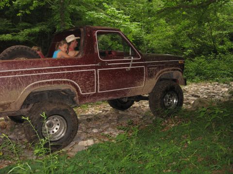 1980 Ford Bronco - A maroon Ford Bronco navigates a rough, wooded trail with two occupants.