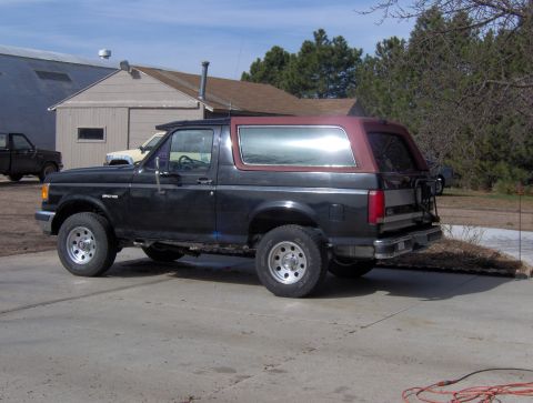 1987 Ford Bronco - A black 1987 Ford Bronco with a maroon top is parked in a driveway next to a building.
