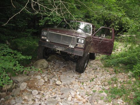 1980 Ford Bronco - This maroon 1980 Ford Bronco is shown off-roading on a rocky, wooded path with its driver's door ajar.