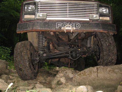 1980 Ford Bronco - The front end of a lifted, dark red Ford Bronco is shown angled down towards rocks and foliage. The vehicle features large off-road tires,…